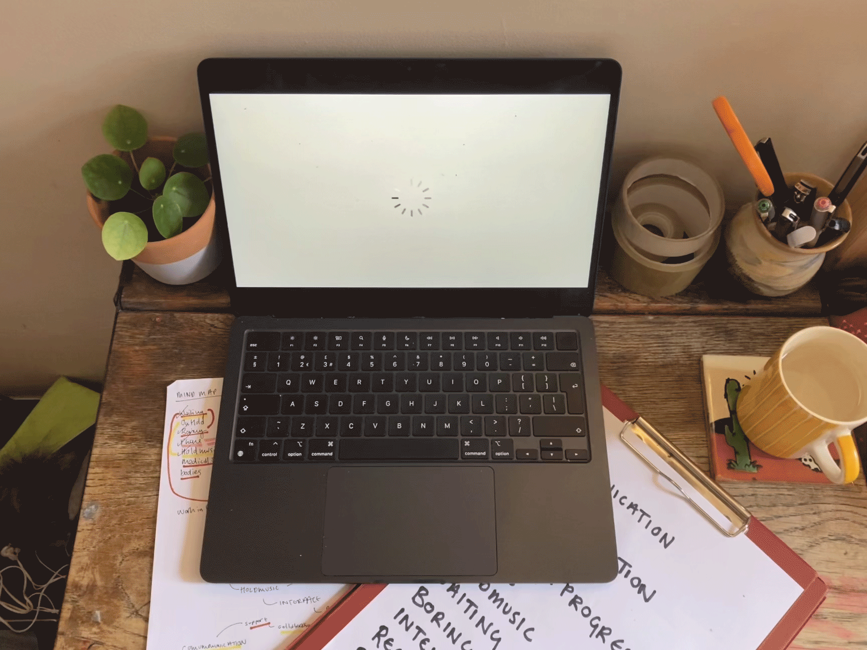 A laptop on a small wooden desk on top of hand written notes. The screen is blank, with a rotating circular loading symbol in the centre and the laptop is surrounded by a pen pot, rolls of tape, a mug and a plant pot.