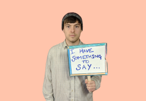 Artist Gary-Martin from the waist up on a light pink background, wearing a pale checked shirt and grey beanie hat with brown hair sticking out. He is holding a sign that reads: ‘I have something to say…’ 