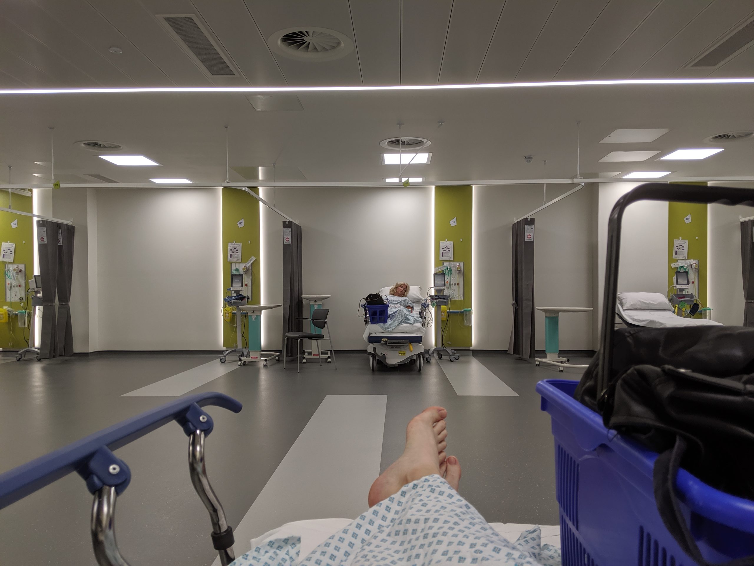 A view of a hospital ward that looks down a bed to a pair of feet and beyond to curtained sections that are completely empty, have a bed with another patient and medical equipment in, and another with medical equipment and an empty bed.
