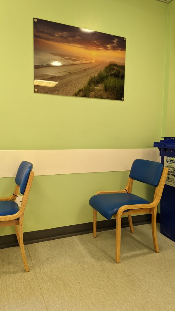 Two chairs in a hospital waiting area in harsh light, next to a pale green wall that has a photo of an expansive sandy beach at sunset hanging on it. Two ceiling lights are reflected in the picture.