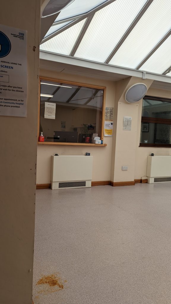 An empty hospital corridor with scuffed well worn floors, beige walls, clunky radiators, and an empty reception area behind a tinted window. 