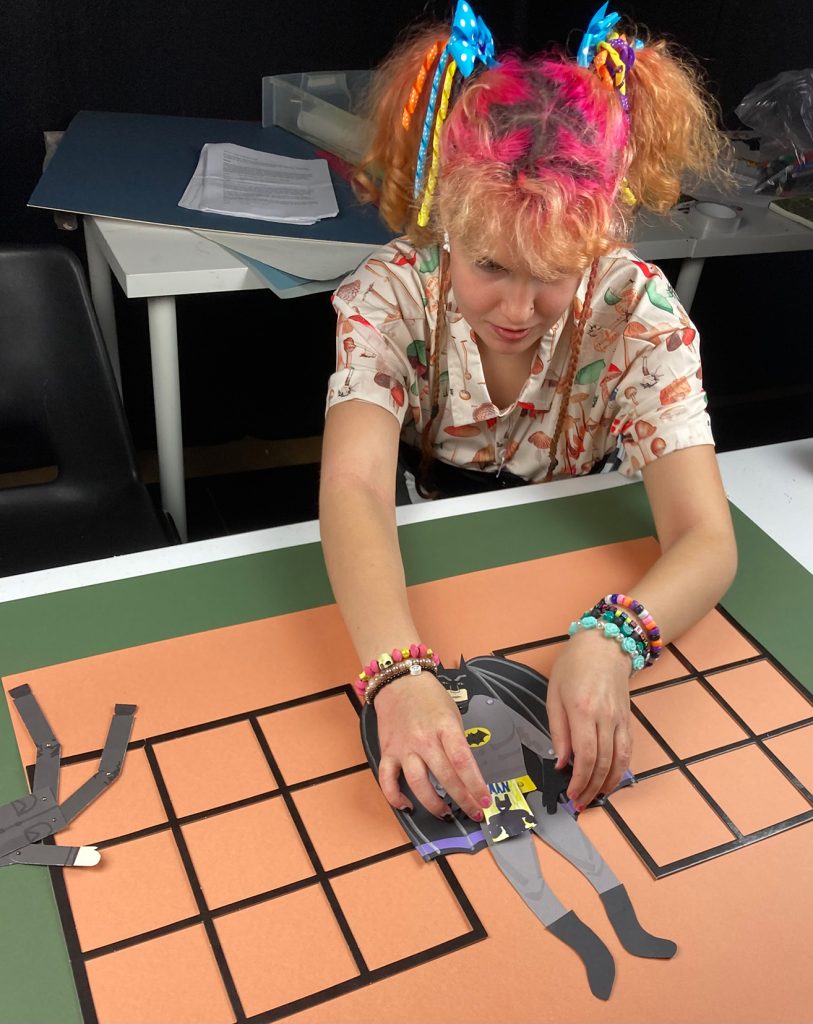 Assistant Keira Hall in a shirt with a mushroom pattern, lots of bracelets, and pink/orange dyed hair in pigtails, reaching across a table to move a paper puppet of Batman on an orange paper background with black grid windows.