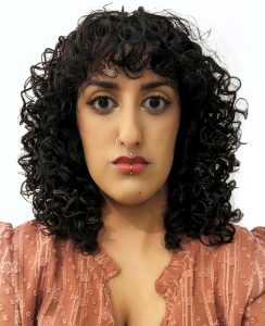 Artist Aminder Virdee from the shoulders up, wearing a terracotta shirt with textured pattern, shoulder length black curly hair with nose and lip piercings.