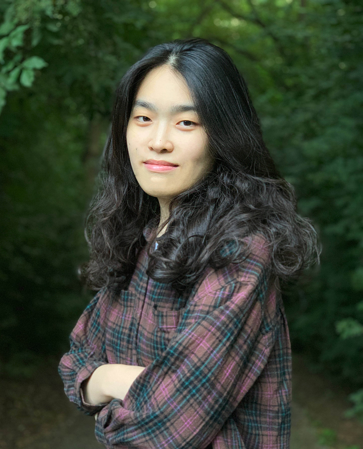 Portrait of the artist Soyhun Lee, with long black hair that becomes wavy at the ends, positioned side on, mouth closed but smiling, with arms folded and head turned towards the camera, wearing a checked/flannel patterned shirt in front of green foliage.